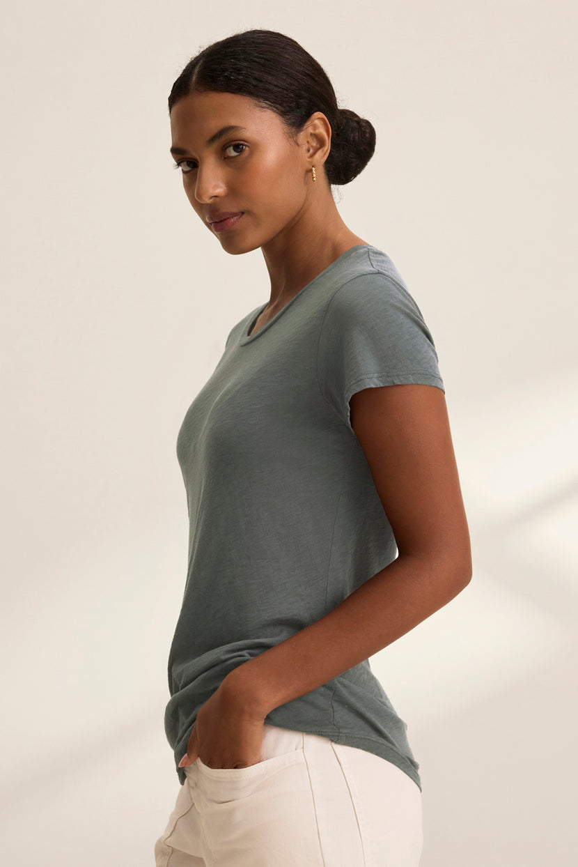 A woman wearing the ODELIA TEE by Velvet by Graham & Spencer, crafted from green cotton slub fabric, pairs it with beige pants. She stands against a light background, hands in pockets, looking sideways. This versatile tee is a true wardrobe essential.