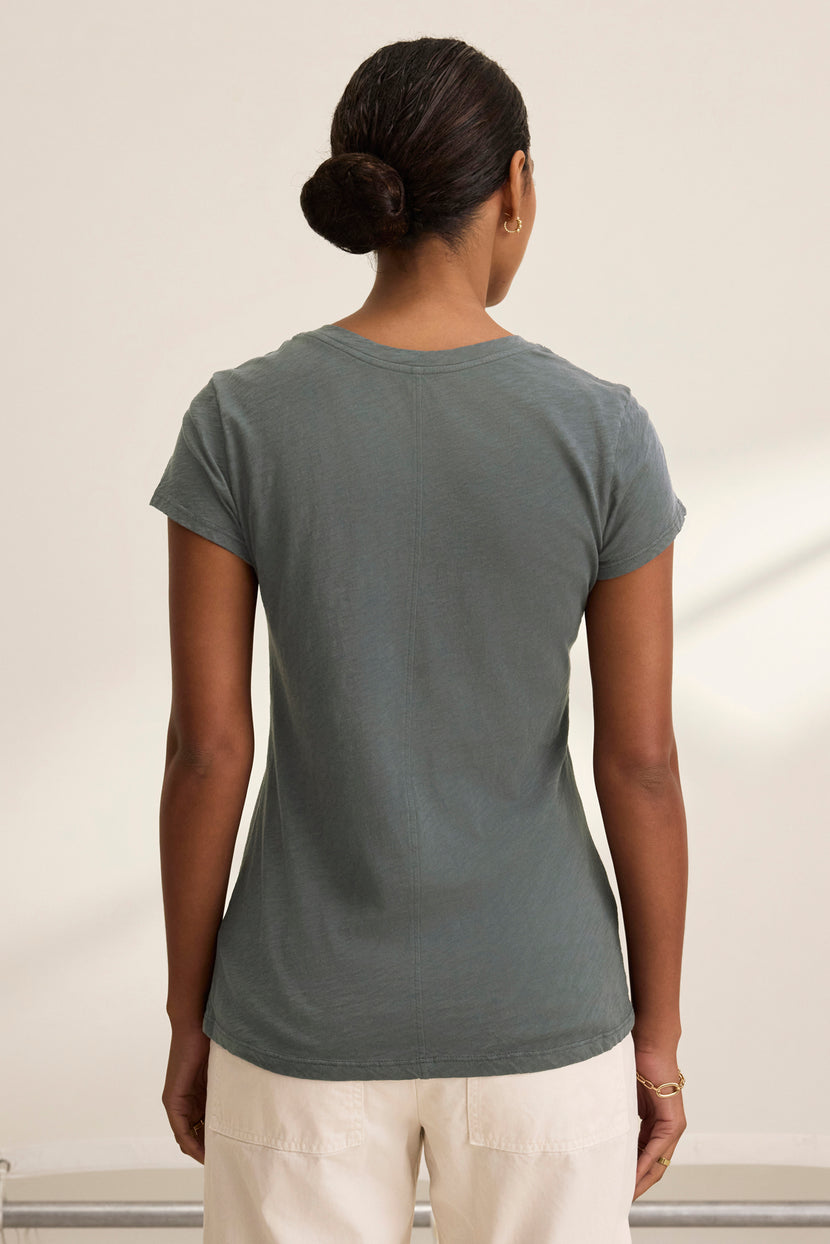 Rear view of a person with a bun hairstyle, wearing the dark green ODELIA TEE by Velvet by Graham & Spencer made from cotton slub fabric paired with white pants, standing against a plain background.