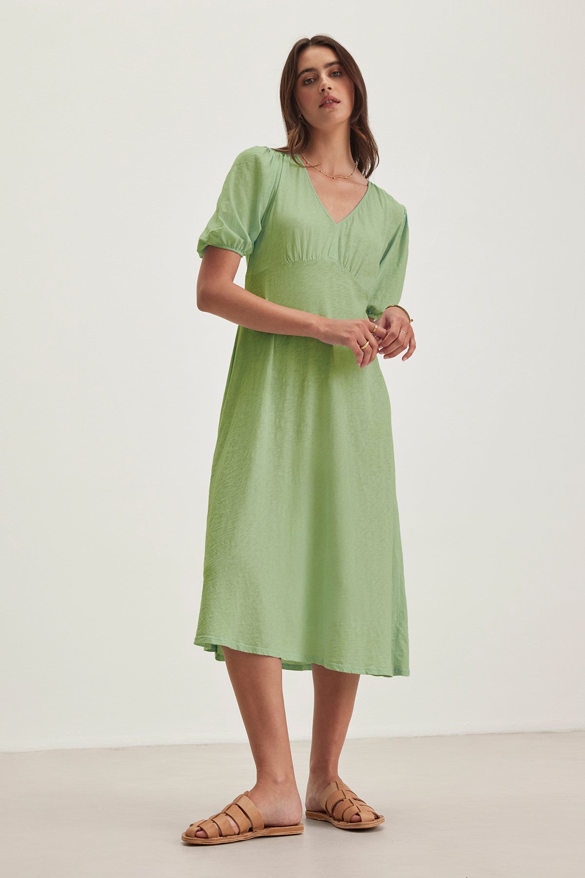   Barefoot in her light green PARKER DRESS by Velvet by Graham & Spencer, a woman stands against a plain white background and light-colored floor. The figure-skimming cotton slub dress enhances her effortless style. 