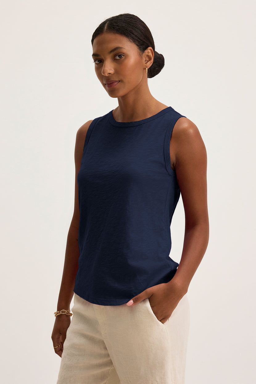 Wearing the TAURUS TANK TOP from Velvet by Graham & Spencer, a person stands with one hand in their pocket, showcasing effortless style in this navy sleeveless top made from textured cotton slub and beige pants.