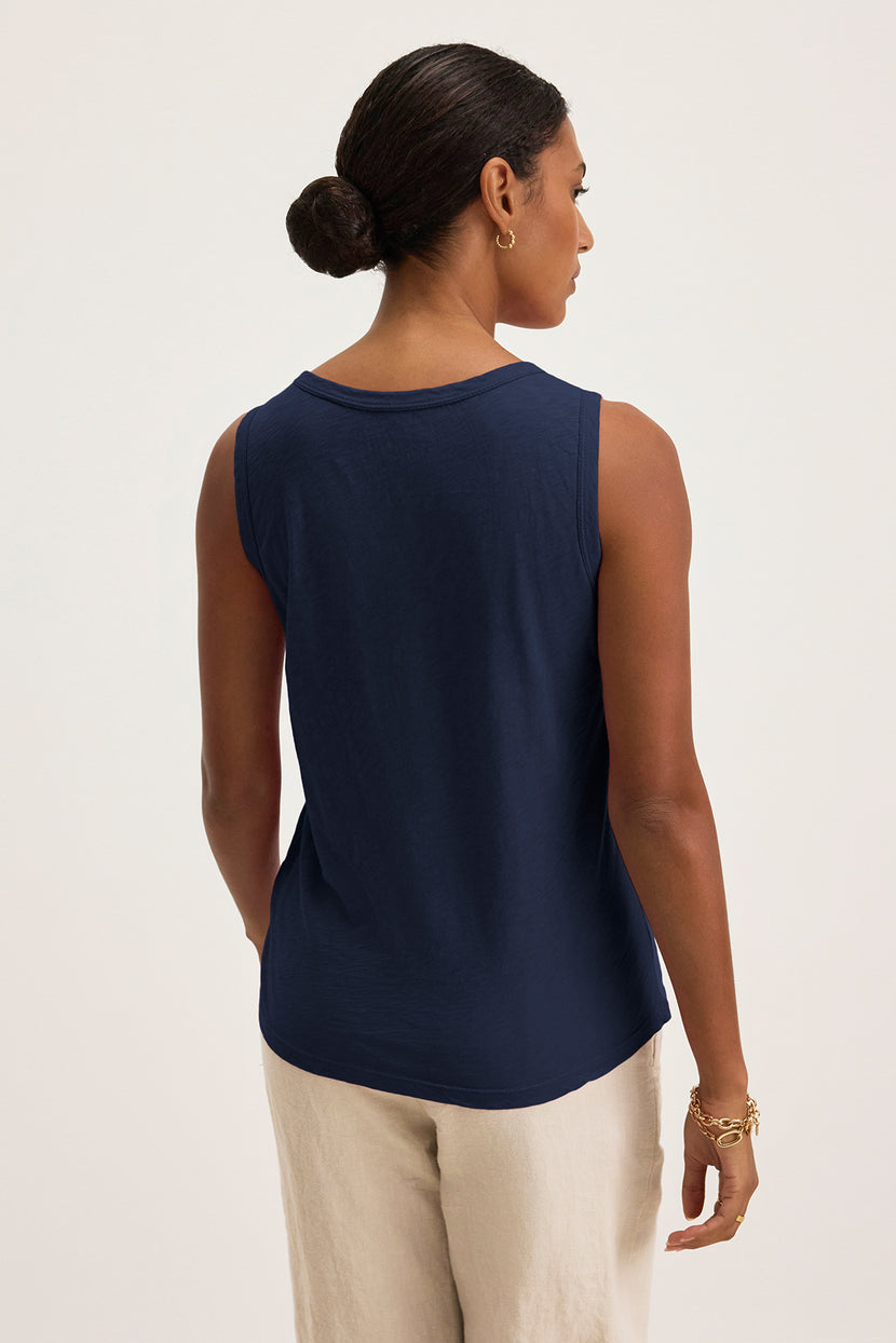 A woman with her hair in a bun is seen from behind wearing the Velvet by Graham & Spencer's essential TAURUS TANK TOP, a navy sleeveless piece crafted from textured cotton slub, paired with beige pants.