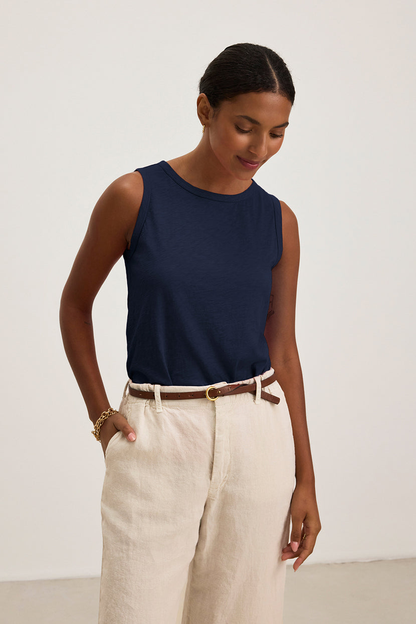 A person wears a navy TAURUS TANK TOP by Velvet by Graham & Spencer with beige pants, looking down and hands in pockets against a plain background. The essential piece features textured cotton slub for subtle interest to the classic look.