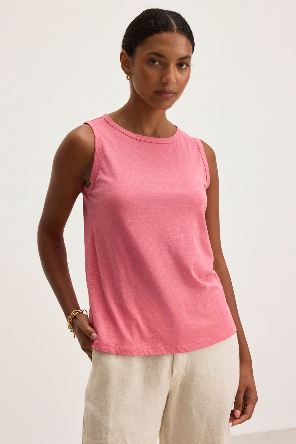   A person models the TAURUS TANK TOP by Velvet by Graham & Spencer, a sleeveless pink cotton slub piece, against a plain background, highlighting its essential crew neck design alongside light-colored pants. 