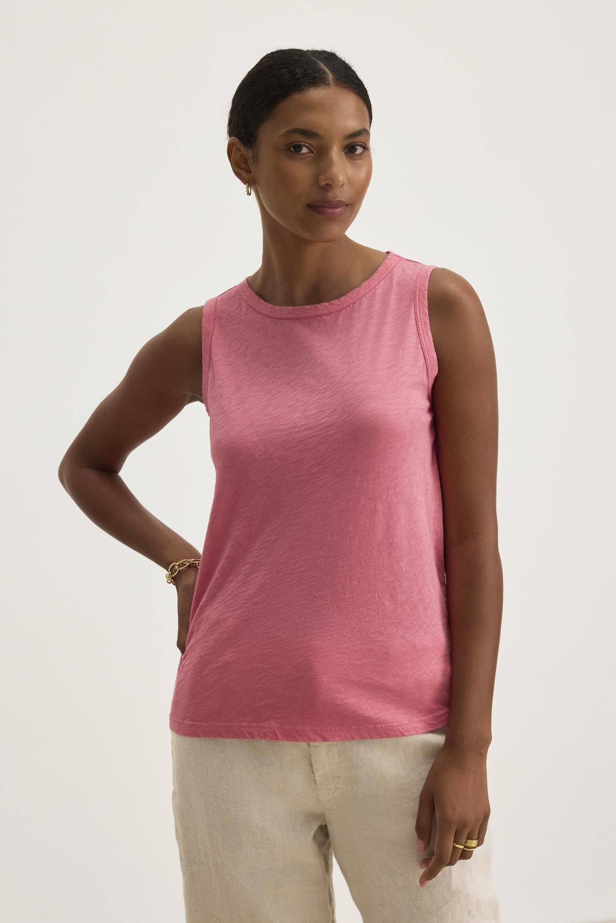   Someone wearing the TAURUS TANK TOP by Velvet by Graham & Spencer, a pink sleeveless essential in textured cotton slub, pairs it with beige pants against a plain background. 