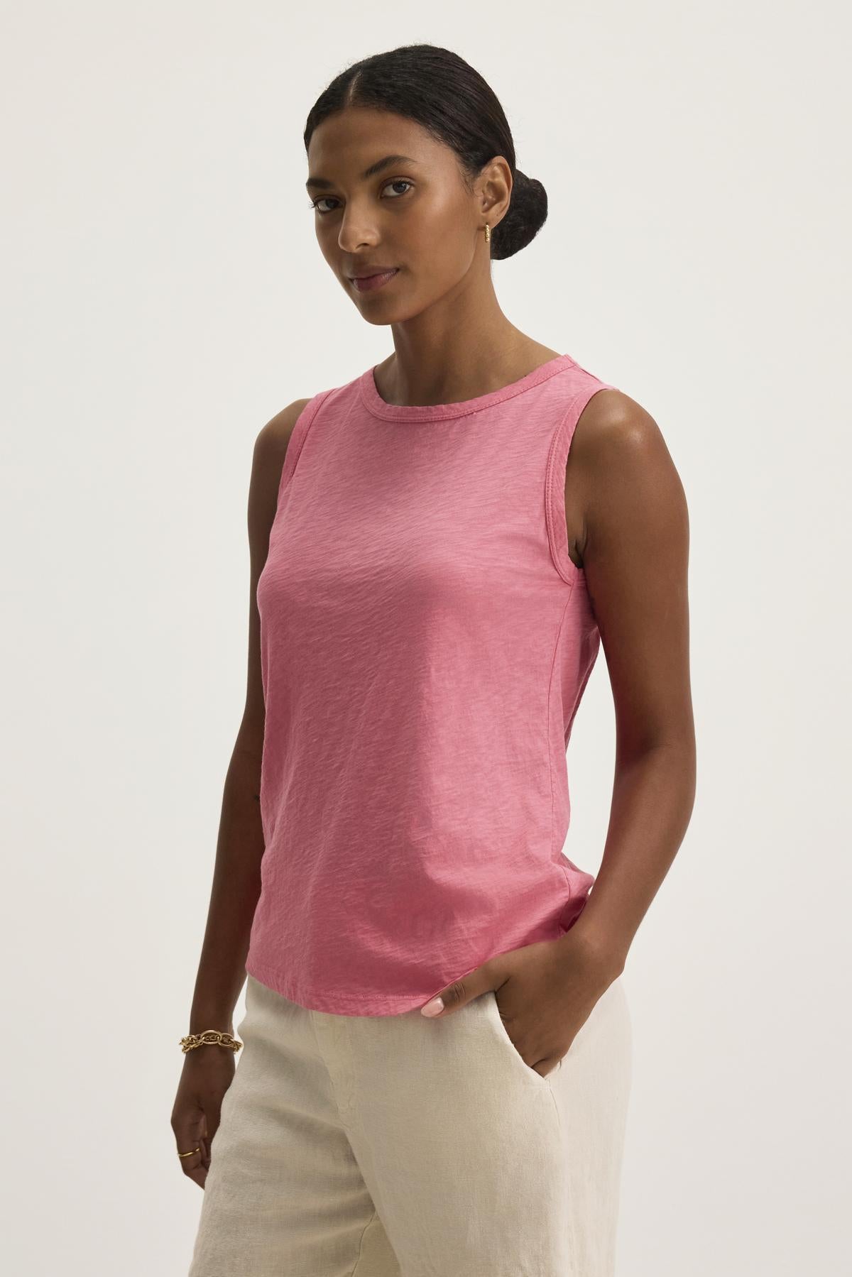   A person wearing the TAURUS TANK TOP, a pink sleeveless top by Velvet by Graham & Spencer, stands against a plain background with light-colored pants and one hand casually in their pocket, effortlessly balancing style and comfort with its timeless crew neck design. 