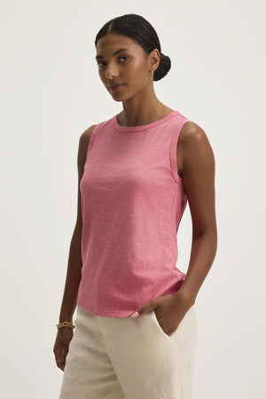 A person wearing the TAURUS TANK TOP, a pink sleeveless top by Velvet by Graham & Spencer, stands against a plain background with light-colored pants and one hand casually in their pocket, effortlessly balancing style and comfort with its timeless crew neck design.