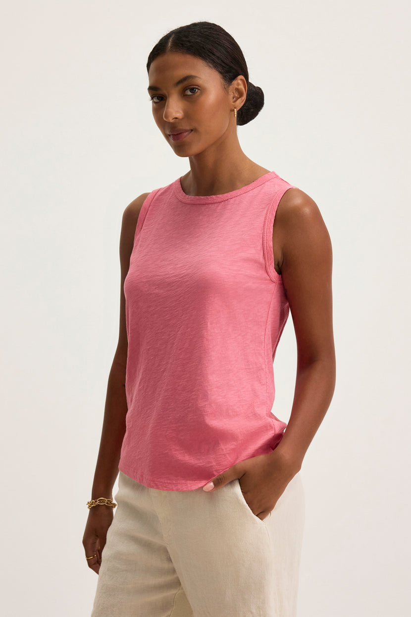 A person wearing the TAURUS TANK TOP, a pink sleeveless top by Velvet by Graham & Spencer, stands against a plain background with light-colored pants and one hand casually in their pocket, effortlessly balancing style and comfort with its timeless crew neck design.