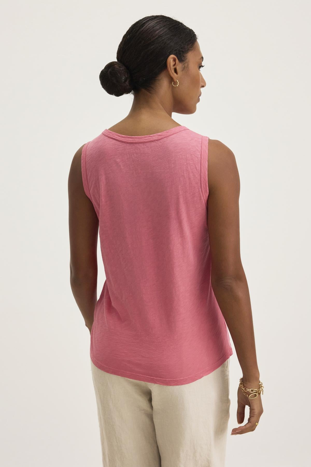   A person in a pink Taurus Tank Top by Velvet by Graham & Spencer, an essential piece with a timeless crew neck design, paired with beige pants, faces away with hair in a bun. 