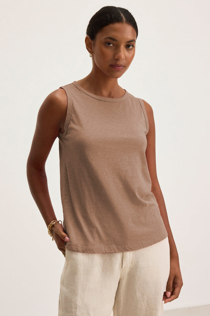 A woman wears the TAURUS TANK TOP by Velvet by Graham & Spencer—a sleeveless taupe crew neck style—paired with light beige pants against a plain background, showcasing an essential piece for effortless style.