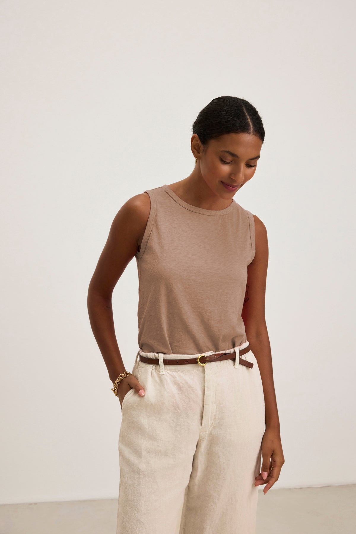   A woman stands against a light background, wearing the TAURUS TANK TOP by Velvet by Graham & Spencer—a sleeveless beige textured cotton slub top with a crew neck—paired with white high-waisted pants, a brown belt, and a gold bracelet, hands in pockets. 