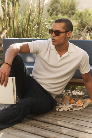 Wearing the DILLON SWEATER POLO by Velvet by Graham & Spencer, a man in sunglasses relaxes on a wooden deck with a closed laptop and a net bag of oranges beside him, enjoying this perfect warm-weather layer.