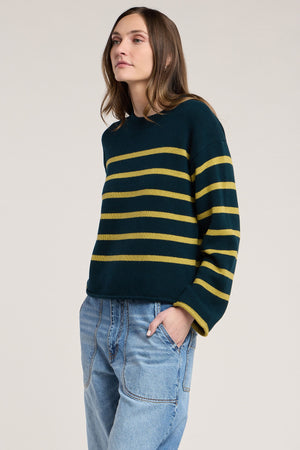 Model wears the LUCIA SWEATER by Velvet by Graham & Spencer—a dark green, relaxed-fit style featuring yellow horizontal stripes and a classic crew neckline—paired with light blue jeans, standing with one hand in a pocket against a plain background.