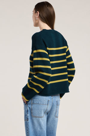 A person wearing the LUCIA SWEATER by Velvet by Graham & Spencer—a dark green, relaxed-fit cotton-cashmere crewneck with yellow horizontal stripes—paired with light blue jeans stands with their back to the camera against a plain background. The sweater features a classic crew neckline, offering a comfortable and timeless look.
