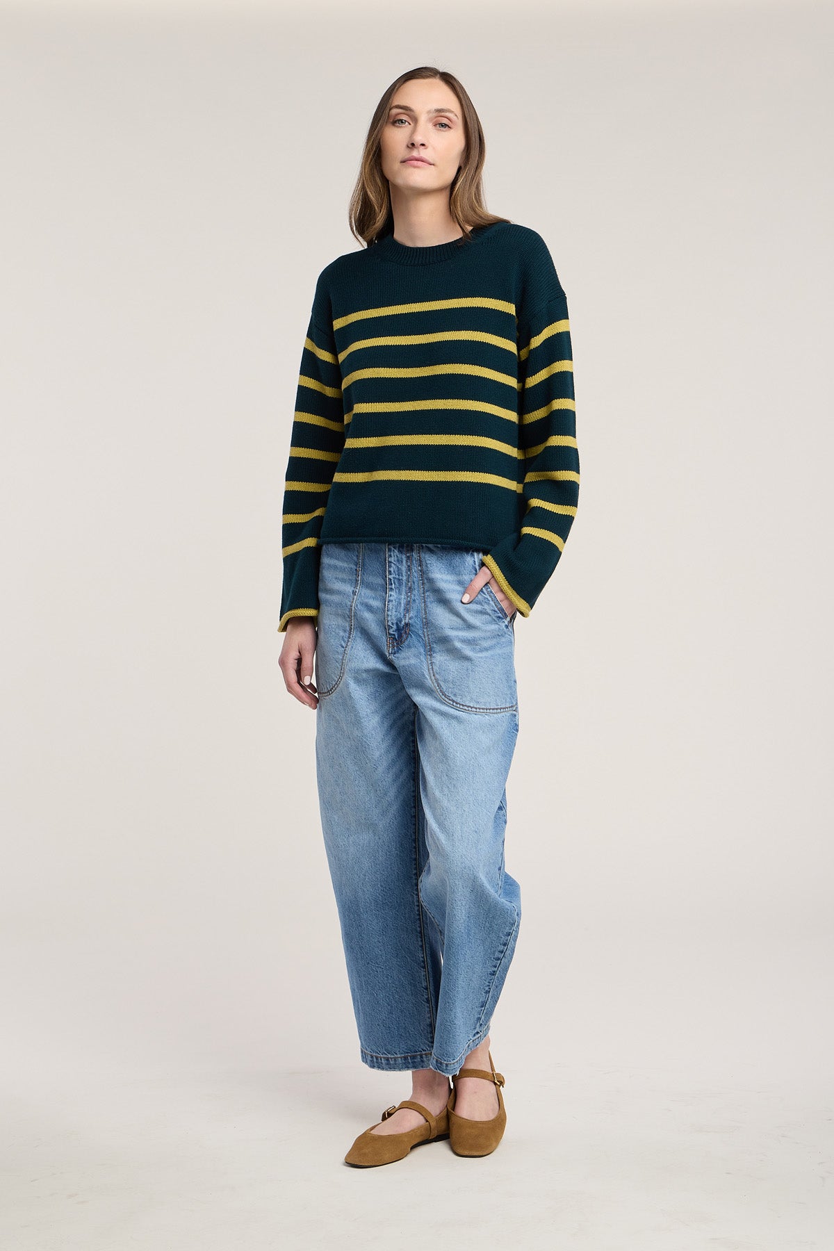 A person stands against a plain background, wearing the relaxed-fit LUCIA SWEATER by Velvet by Graham & Spencer—a green and yellow striped piece with a classic crew neckline—paired with loose blue jeans and brown shoes.