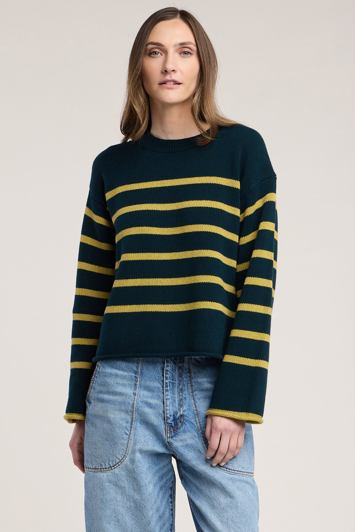A woman stands against a plain background wearing the Velvet by Graham & Spencer LUCIA SWEATER—a dark green, relaxed-fit knit with yellow horizontal stripes and a classic crew neckline—paired with light blue jeans.
