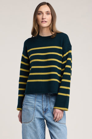 A woman stands against a plain background wearing the Velvet by Graham & Spencer LUCIA SWEATER—a dark green, relaxed-fit knit with yellow horizontal stripes and a classic crew neckline—paired with light blue jeans.