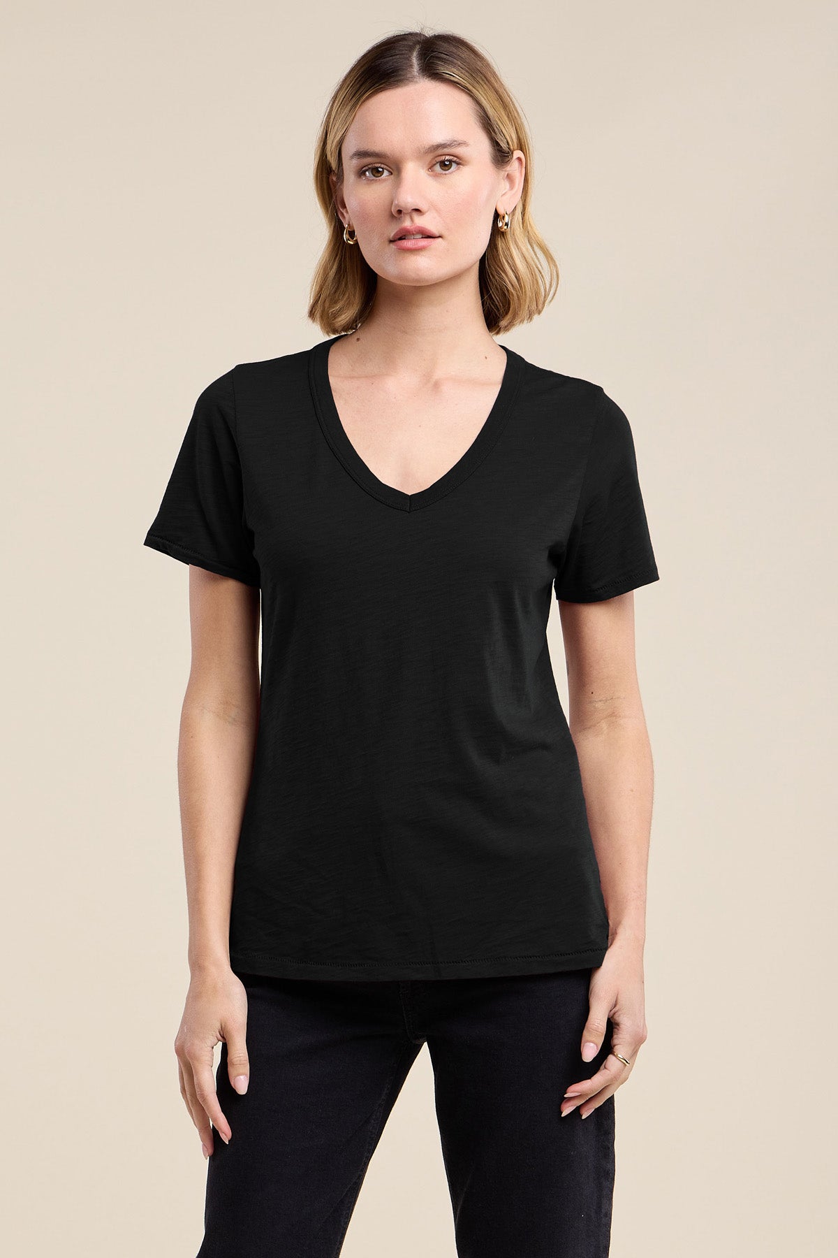A woman faces forward in the Velvet by Graham & Spencer LILITH TEE, a flattering plain black V-neck, paired with black pants against a neutral background.