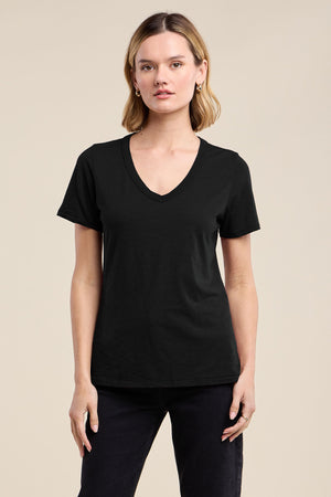A woman faces forward in the Velvet by Graham & Spencer LILITH TEE, a flattering plain black V-neck, paired with black pants against a neutral background.