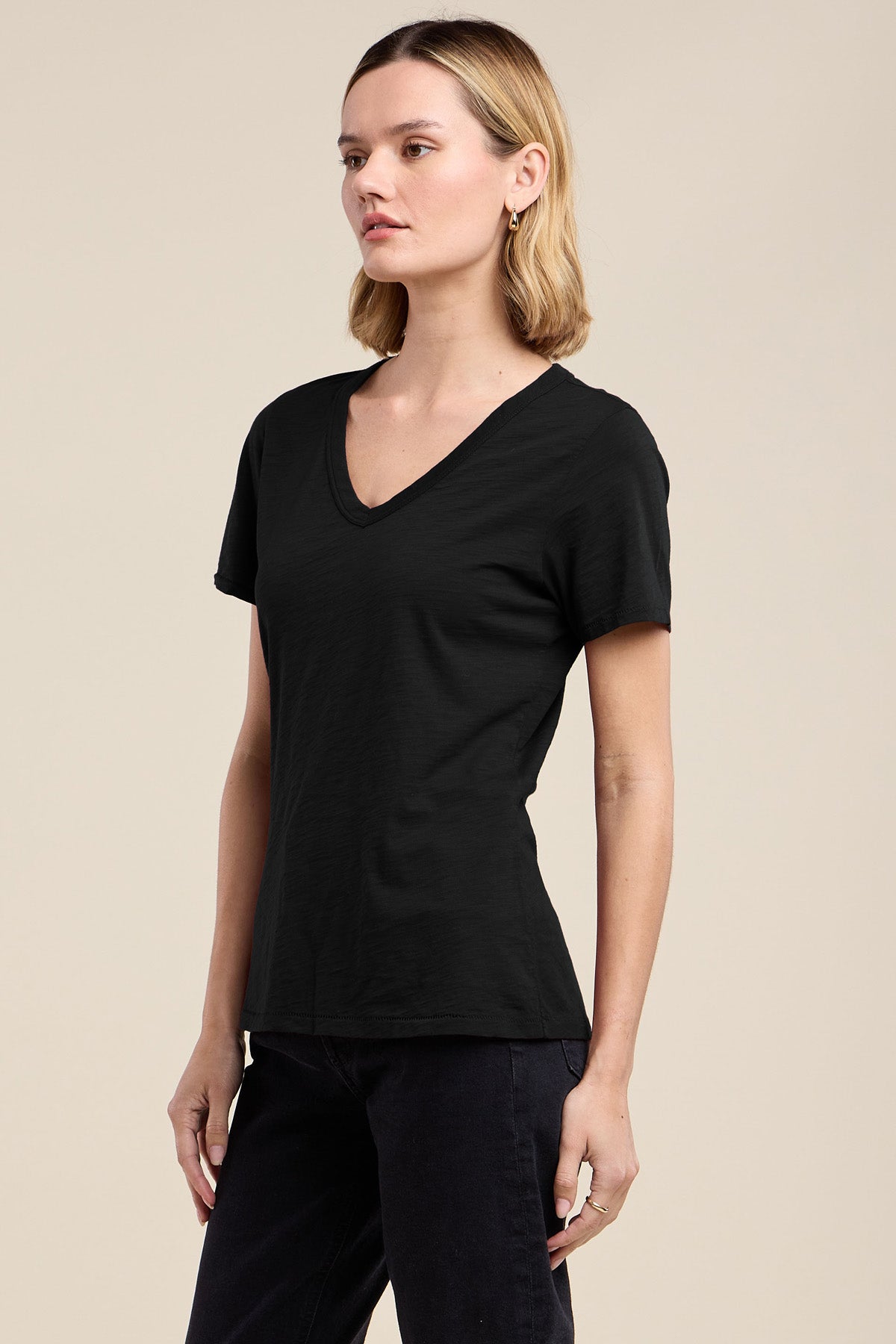 A person with shoulder-length hair wears the LILITH TEE by Velvet by Graham & Spencer, a plain black cotton slub V-neck, paired with black pants and standing against a neutral background.