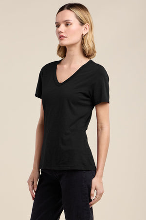 A person with shoulder-length hair wears the LILITH TEE by Velvet by Graham & Spencer, a plain black cotton slub V-neck, paired with black pants and standing against a neutral background.
