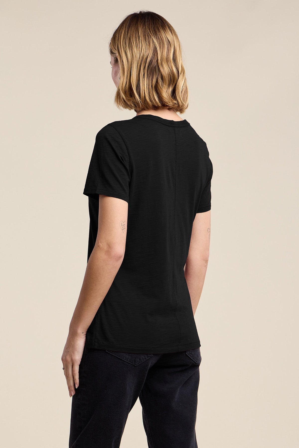 Person with short, wavy hair wears the LILITH TEE by Velvet by Graham & Spencer—a flattering black V-neck—paired with dark pants. They're standing with their back to the camera against a plain backdrop.