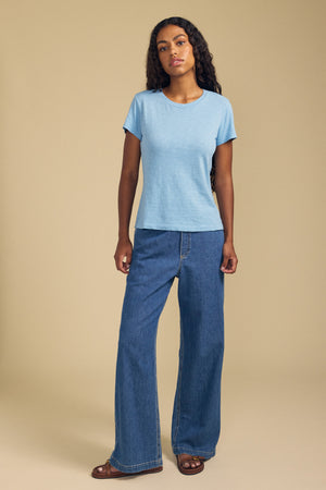 A woman stands against a beige background, wearing the ODELIA TEE by Velvet by Graham & Spencer—a light blue cotton slub top with a classic crew neckline—paired with wide-leg blue jeans and brown sandals.