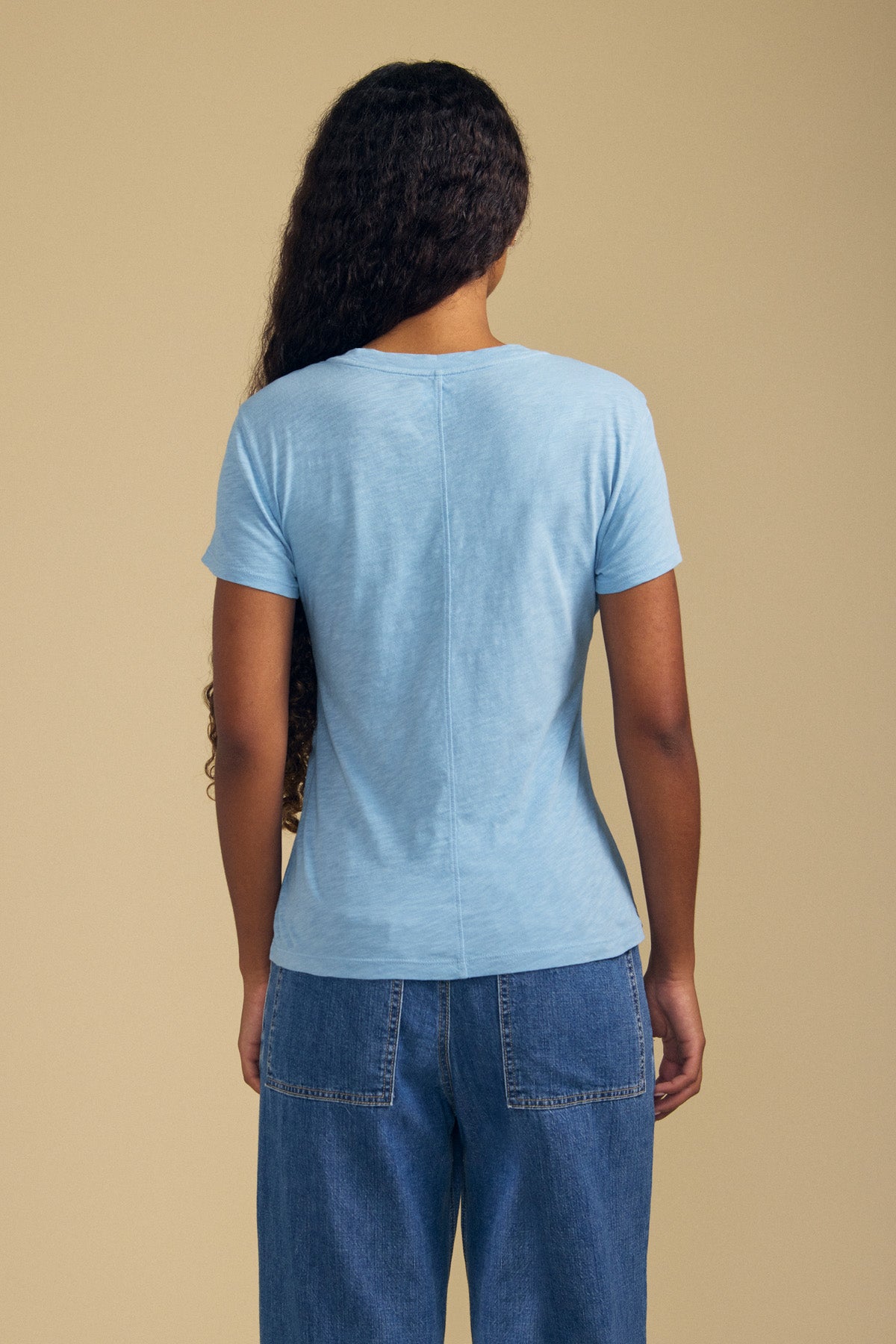 Person with long curly hair, wearing the Velvet by Graham & Spencer ODELIA TEE—a light blue cotton slub tee with a classic crew neckline—paired with blue jeans and standing facing away against a beige background; a true wardrobe essential.-41779747946689
