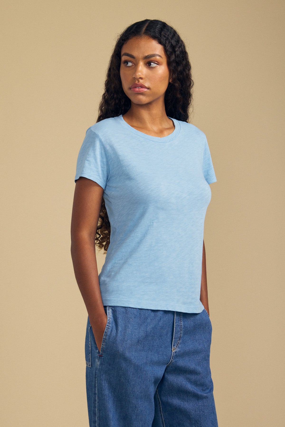A woman with long curly hair wears the Velvet by Graham & Spencer ODELIA TEE—a light blue cotton slub top with a classic crew neckline—paired with blue jeans, standing against a beige background with her hands in her pockets and looking to the side, showcasing this true wardrobe essential.-41779747848385