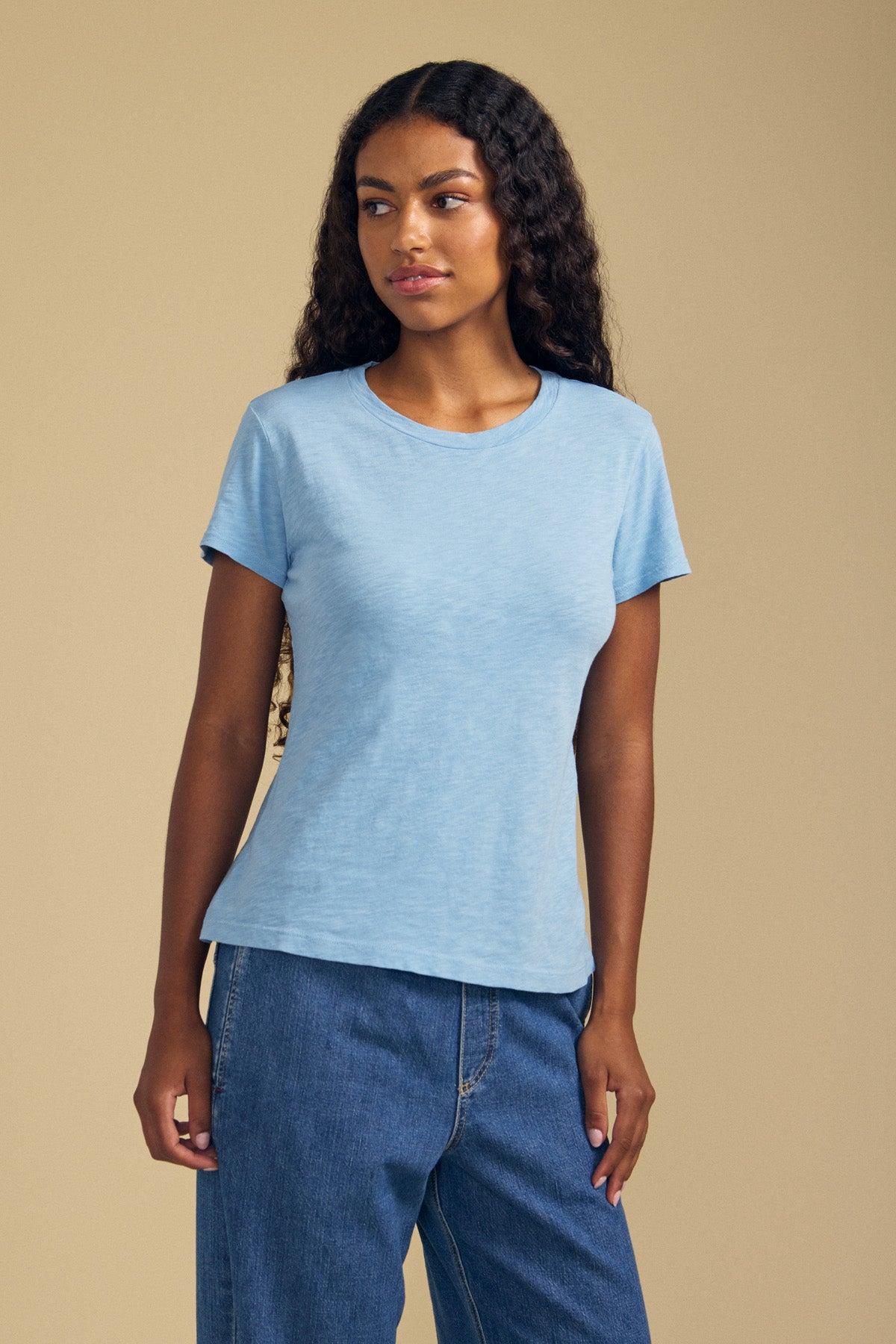 A woman with long curly hair wearing the ODELIA TEE by Velvet by Graham & Spencer—a light blue cotton slub tee featuring a classic crew neckline—paired with blue jeans, stands against a plain beige background, showcasing this true wardrobe essential.-41779747815617