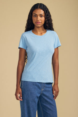 A person with long curly hair wears the ODELIA TEE by Velvet by Graham & Spencer—a light blue premium cotton top with a classic crew neckline—paired with blue jeans, standing against a plain beige background.