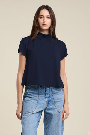 A woman stands against a plain background wearing the SAVI TEE by Velvet by Graham & Spencer—a navy short-sleeve relaxed turtleneck top with a boxy silhouette—paired with high-waisted blue jeans.