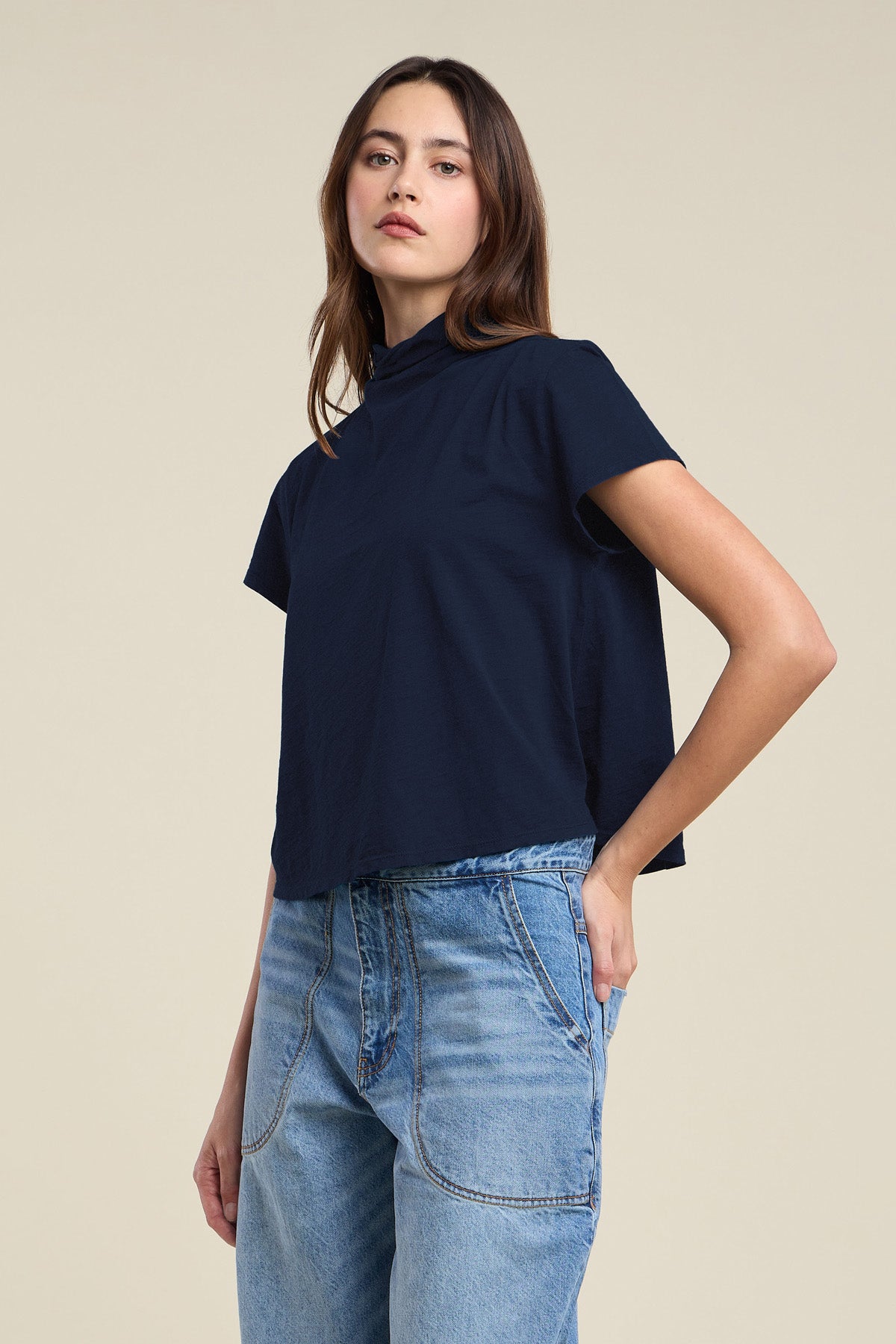 A woman wearing the SAVI TEE by Velvet by Graham & Spencer—a navy blue cotton slub tee with a boxy silhouette—paired with light blue high-waisted jeans, stands against a plain beige background.
