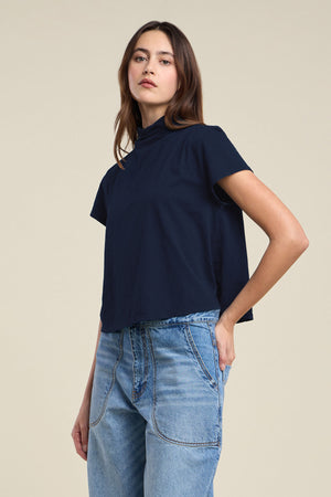 A woman wearing the SAVI TEE by Velvet by Graham & Spencer—a navy blue cotton slub tee with a boxy silhouette—paired with light blue high-waisted jeans, stands against a plain beige background.