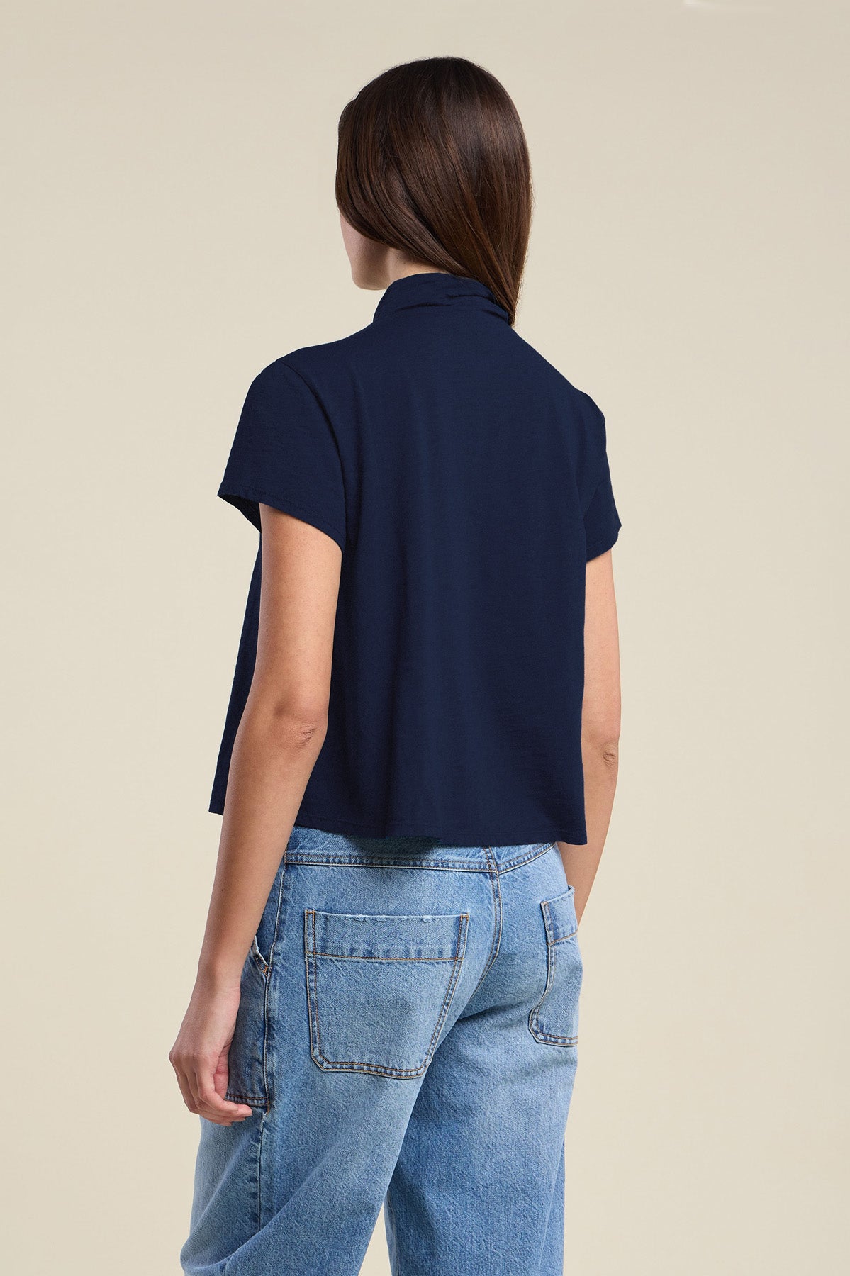 A person stands facing away, wearing the SAVI TEE by Velvet by Graham & Spencer—a navy cotton slub shirt with a boxy silhouette—paired with light blue jeans, against a plain beige background.-42119074939073