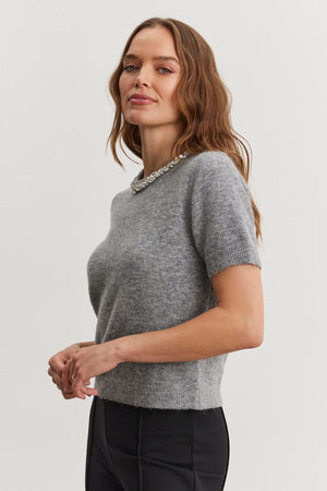 A woman with long hair wears the Velvet by Graham & Spencer LAYTON ALPACA EMBELLISHED SWEATER and black pants, standing against a plain background.