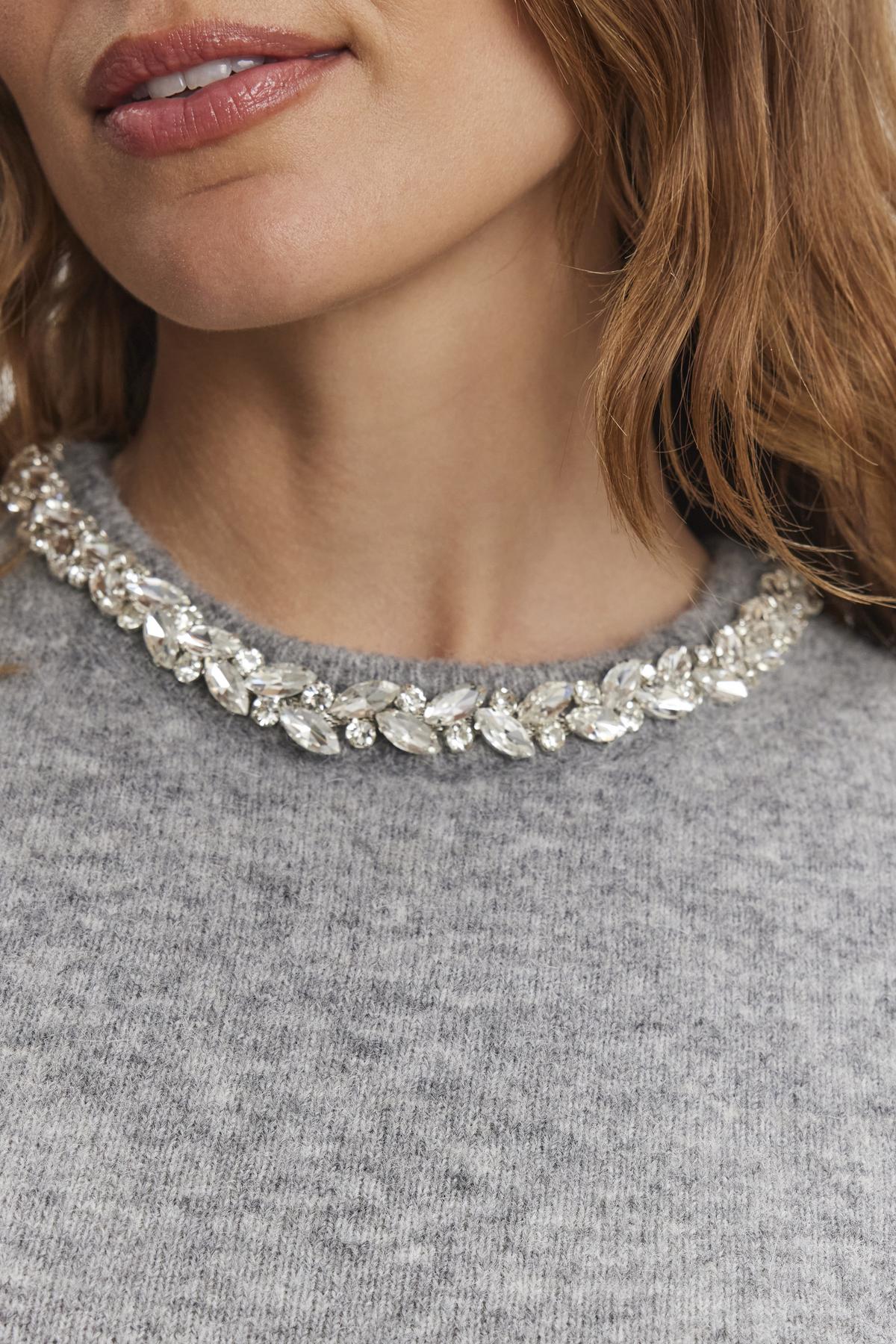   Close-up of a person wearing the LAYTON ALPACA EMBELLISHED SWEATER by Velvet by Graham & Spencer, featuring a rhinestone-adorned neckline. 