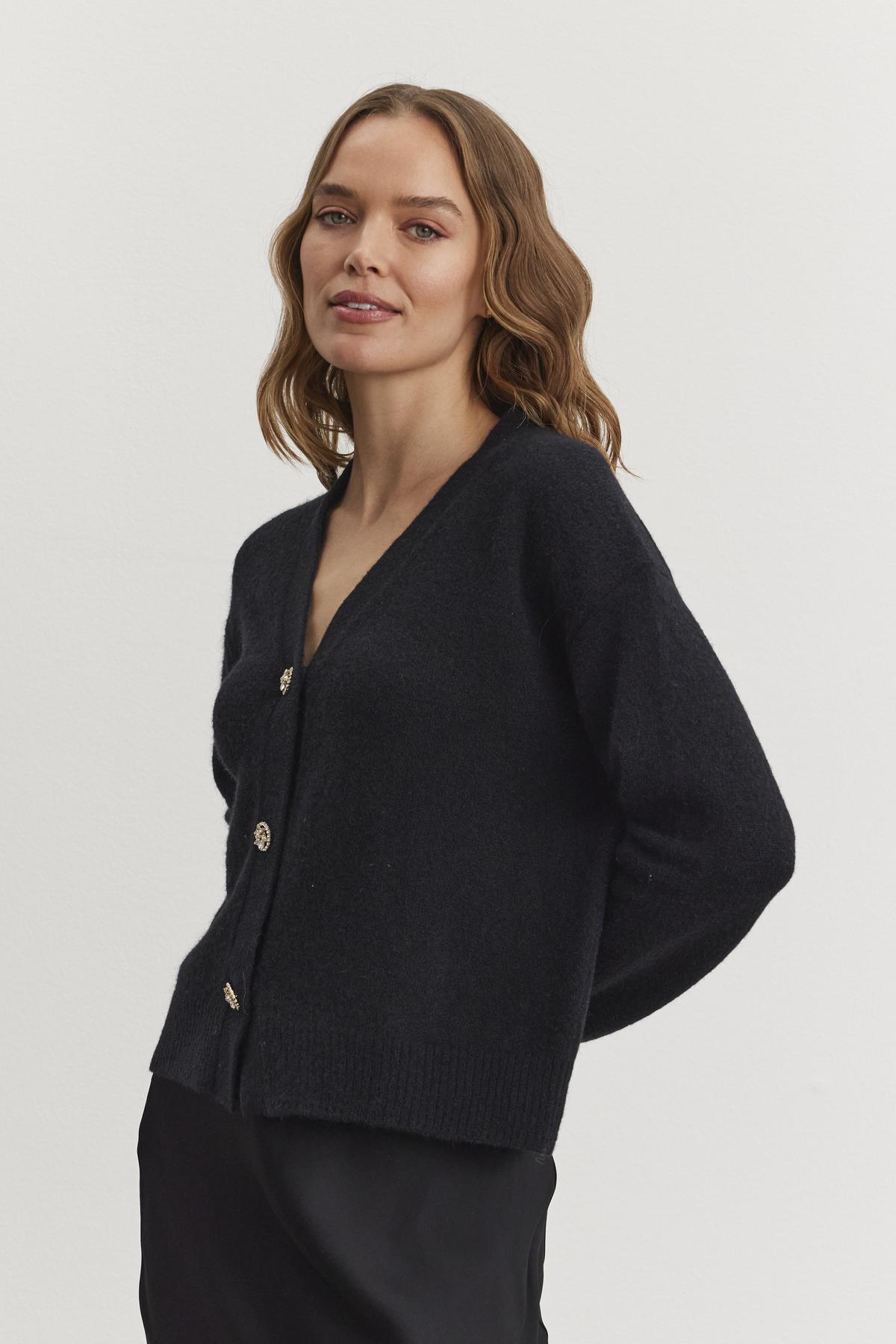   A woman with wavy hair models the MARTI ALPACA EMBELLISHED CARDIGAN by Velvet by Graham & Spencer, paired with a black skirt, as she stands against a plain white background. 