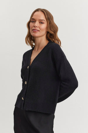 A woman with wavy hair models the MARTI ALPACA EMBELLISHED CARDIGAN by Velvet by Graham & Spencer, paired with a black skirt, as she stands against a plain white background.