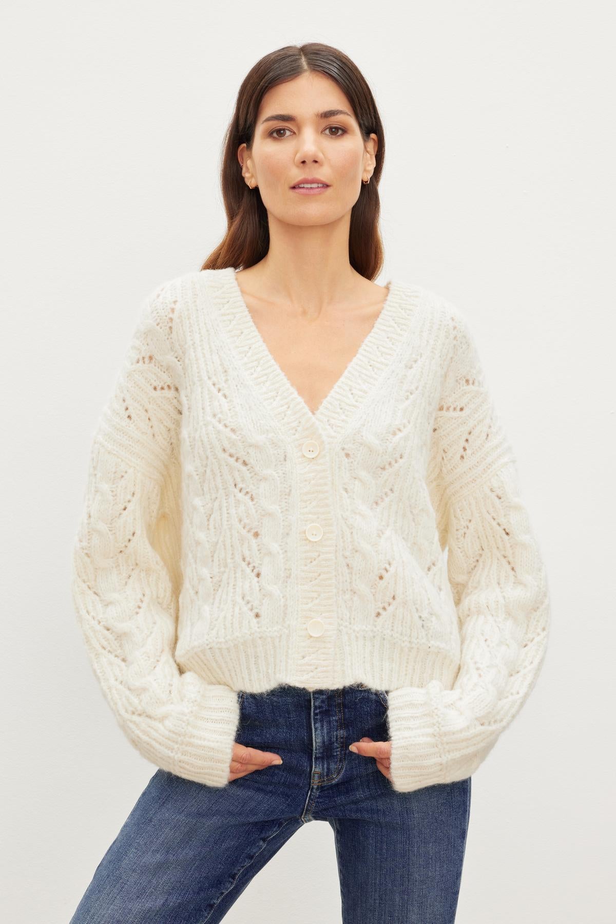   The model is wearing the Velvet by Graham & Spencer ELISA ALPACA BLEND CARDIGAN, a versatile white cable knit sweater with a relaxed fit, paired with jeans. 