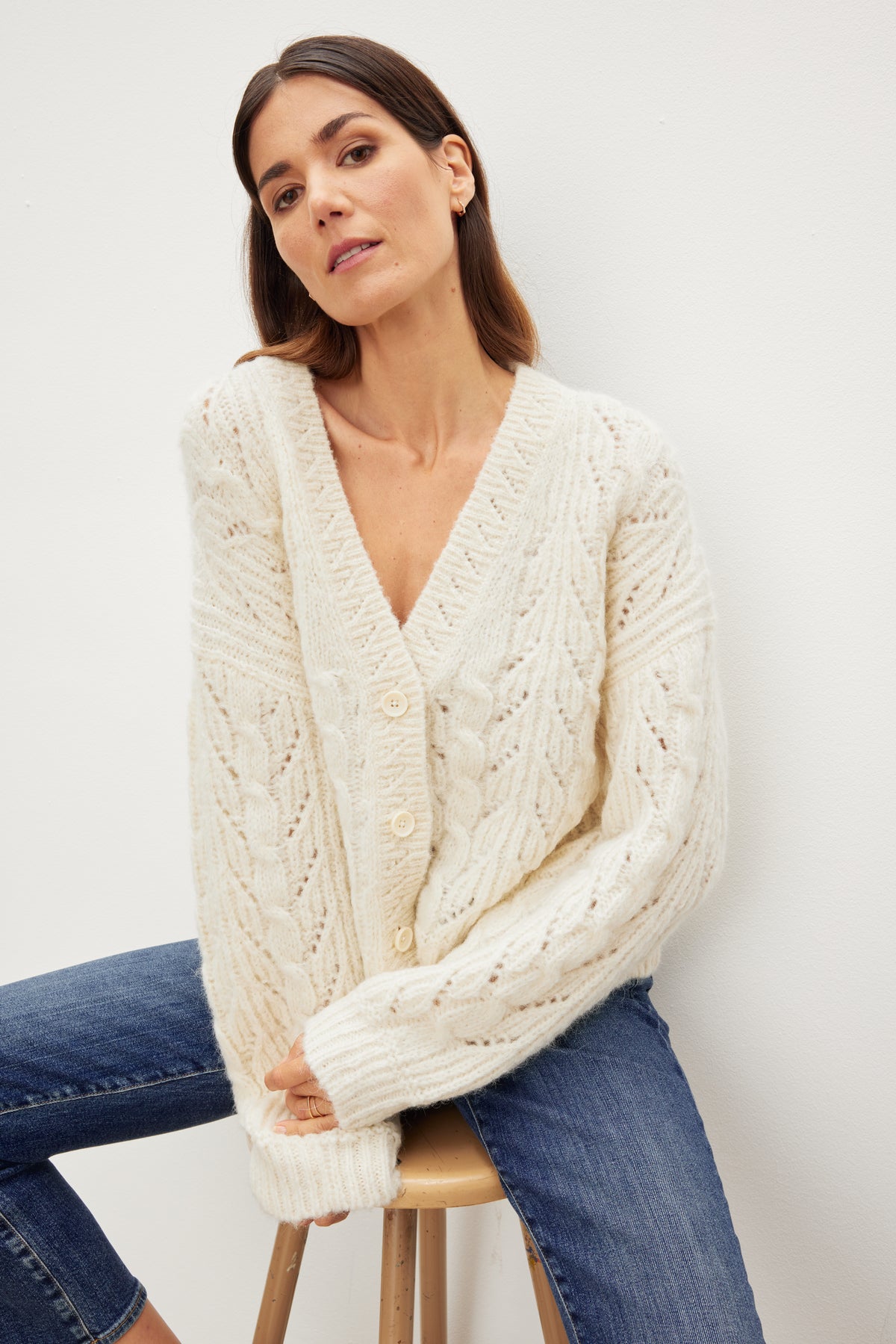 A woman is sitting on a stool wearing relaxed fit jeans and a ELISA ALPACA BLEND CARDIGAN by Velvet by Graham & Spencer.-35655309951169