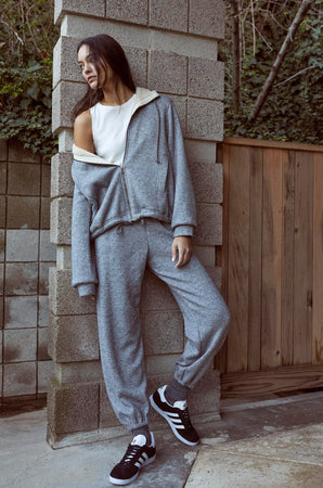 A woman in a grey Velvet by Graham & Spencer sweatsuit leaning against a wall, wearing the OCTAVIA DOUBLE KNIT ZIP UP HOODIE.