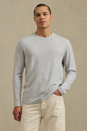 A man wears the COLTON CREW by Velvet by Graham & Spencer—a light gray, long-sleeve crew shirt in soft double-face jersey—with off-white pants, standing against a plain beige background.