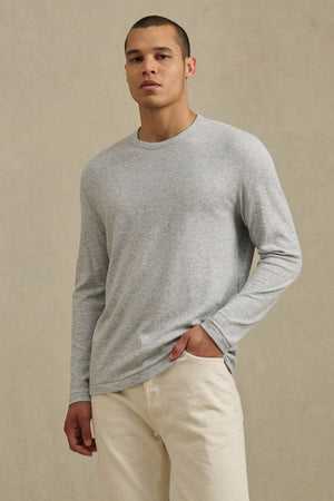 A man wearing the COLTON CREW by Velvet by Graham & Spencer—a light gray long-sleeve crewneck—paired with off-white pants stands against a plain beige background.