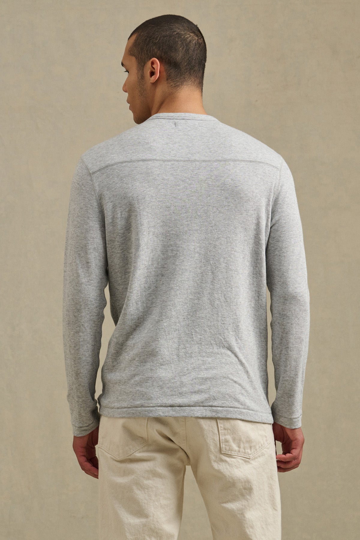 A person stands with their back to the camera, wearing the Velvet by Graham & Spencer COLTON CREW light gray long-sleeve shirt and beige pants with subtle exposed stitching, set against a neutral background.-41008943071425