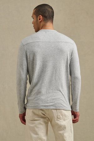 A person stands with their back to the camera, wearing the Velvet by Graham & Spencer COLTON CREW light gray long-sleeve shirt and beige pants with subtle exposed stitching, set against a neutral background.