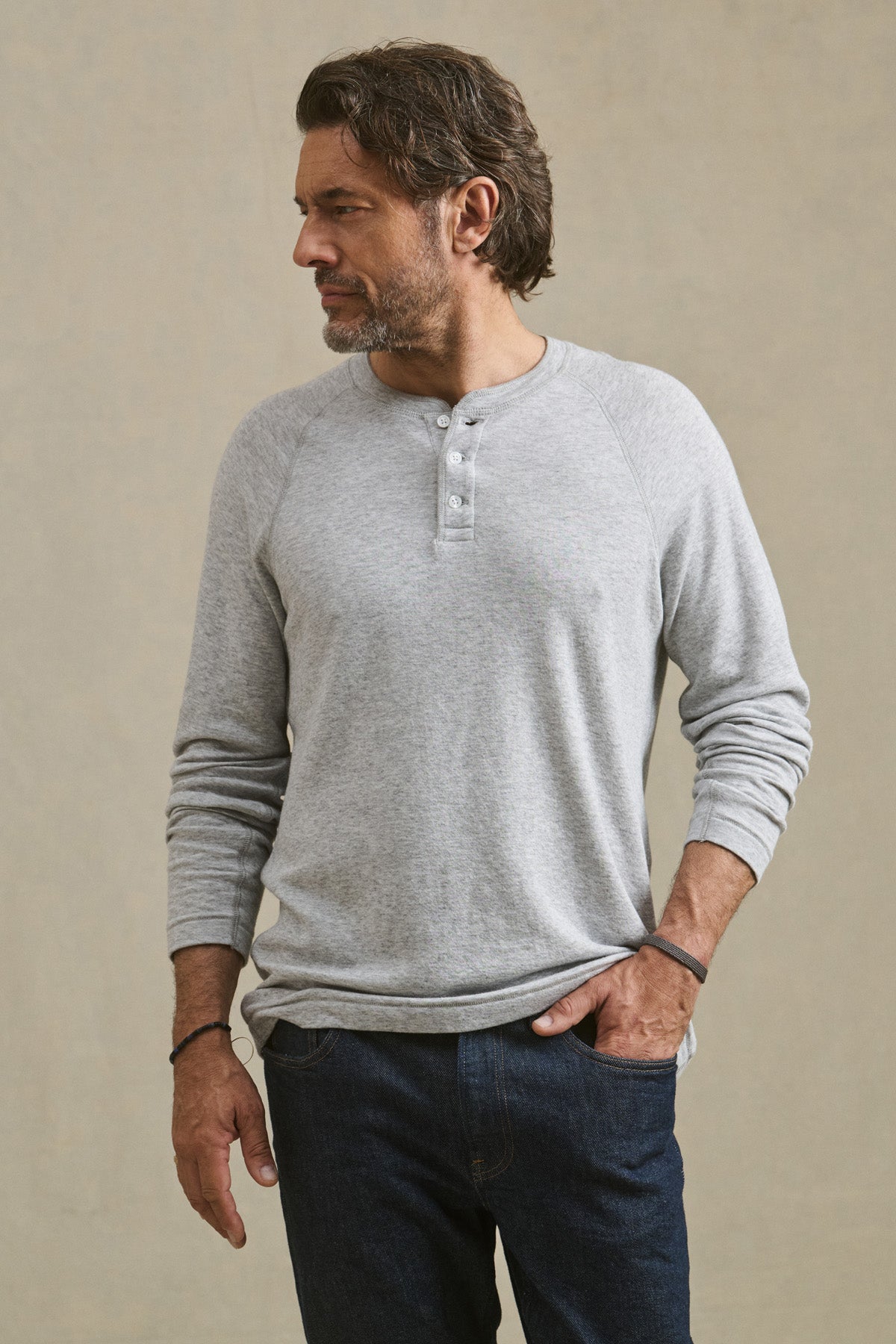 A man with dark hair and a beard wears the WESLEY HENLEY by Velvet by Graham & Spencer in light gray double-face jersey and dark jeans, standing with one hand in his pocket and looking to the side.-41009063952577