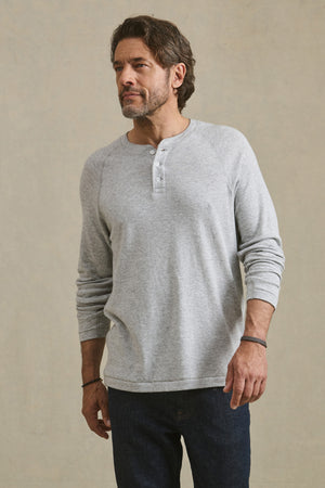 A man wears the Velvet by Graham & Spencer WESLEY HENLEY in light gray and dark blue jeans, standing against a plain beige background.