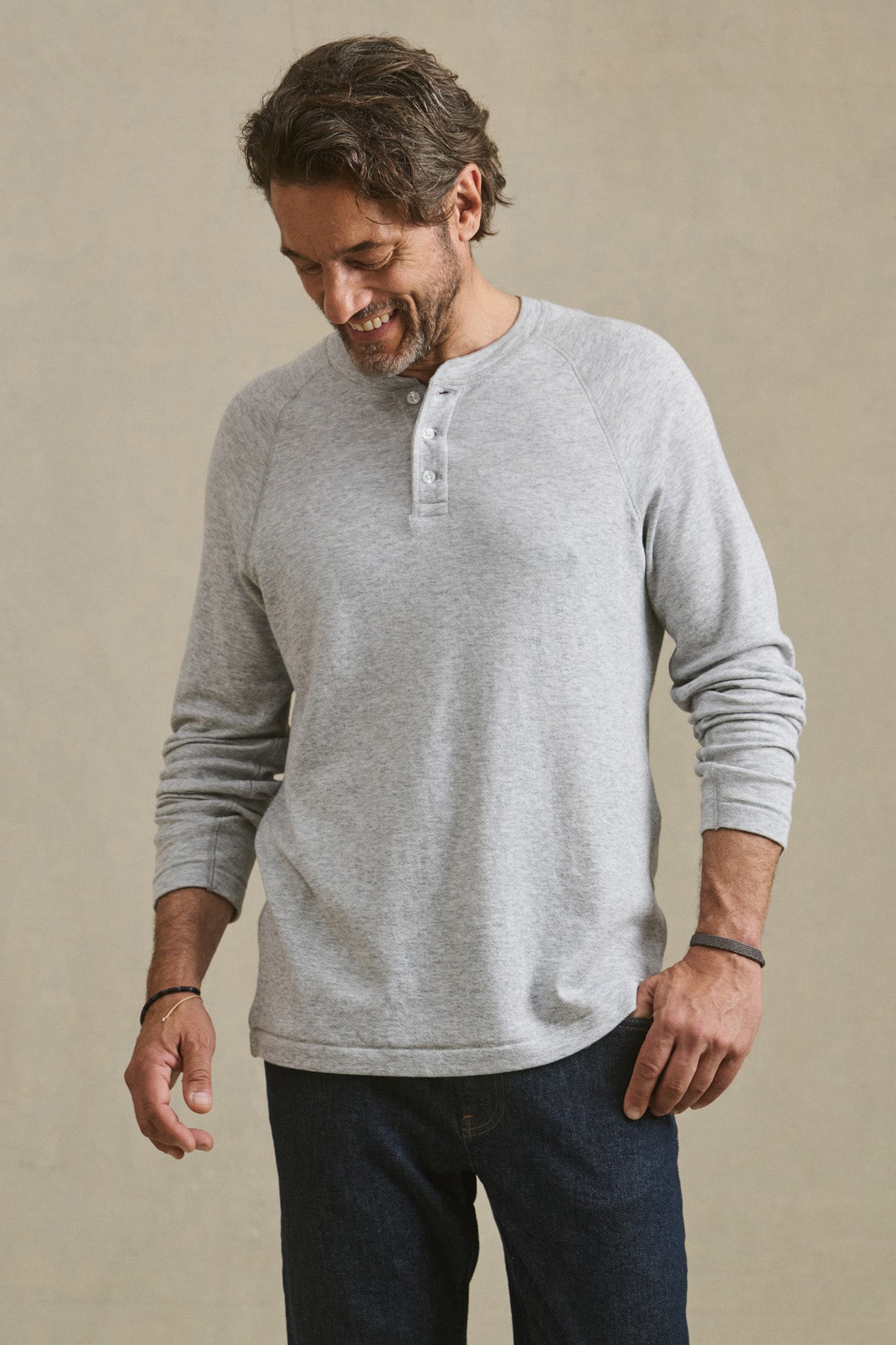 A man wearing the WESLEY HENLEY from Velvet by Graham & Spencer— a light gray long-sleeve men’s casual shirt— pairs it with dark jeans while standing against a plain background, looking down with a slight smile.