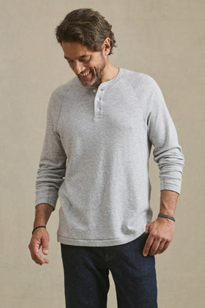 A man wearing the WESLEY HENLEY from Velvet by Graham & Spencer— a light gray long-sleeve men’s casual shirt— pairs it with dark jeans while standing against a plain background, looking down with a slight smile.