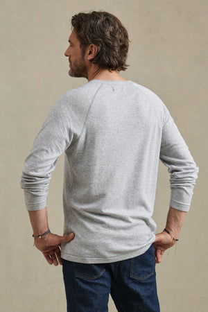 A man faces away from the camera, wearing the WESLEY HENLEY men's tee by Velvet by Graham & Spencer in light gray with dark blue jeans against a plain, neutral background.